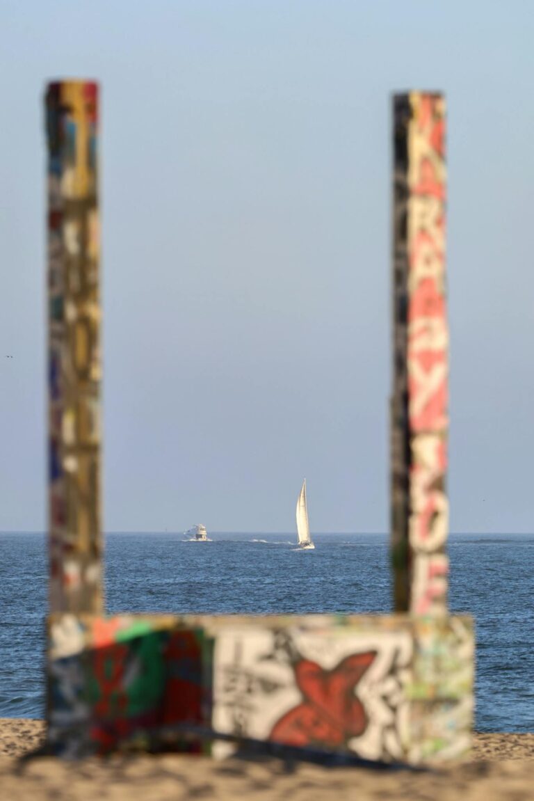 A serene sailboat sailing in the ocean framed by vibrant graffiti-covered poles at a Los Angeles beach.