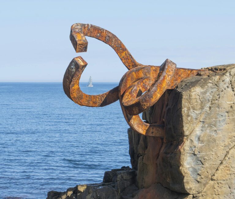 Rusty sculpture by the sea with a distant sailboat in San Sebastián, Spain.