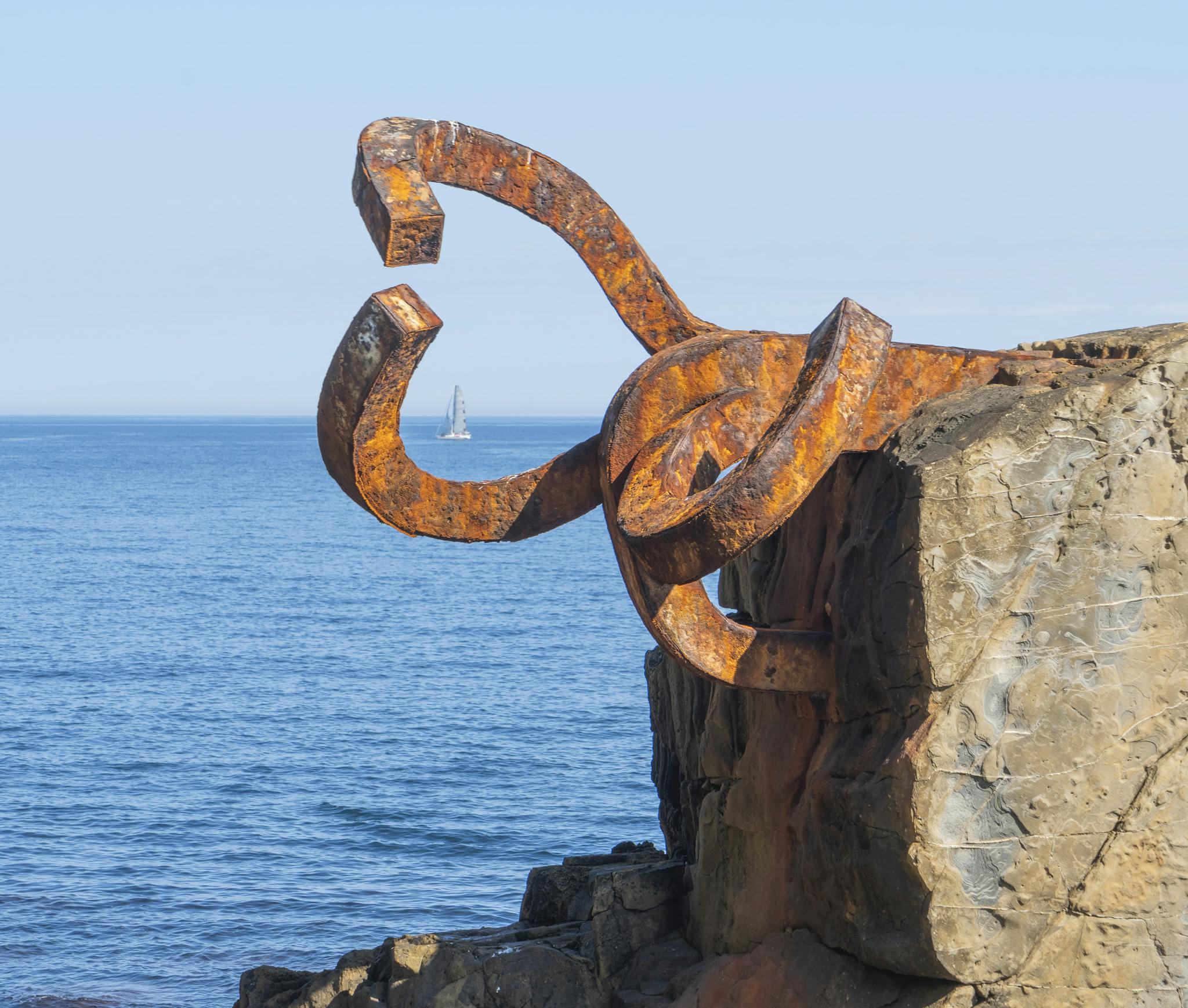 Rusty sculpture by the sea with a distant sailboat in San Sebastián, Spain.